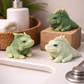 Handmade iguana-shaped goat milk soaps displayed on a bathroom sink and wooden riser with towels and greenery for a spa-inspired lifestyle scene.