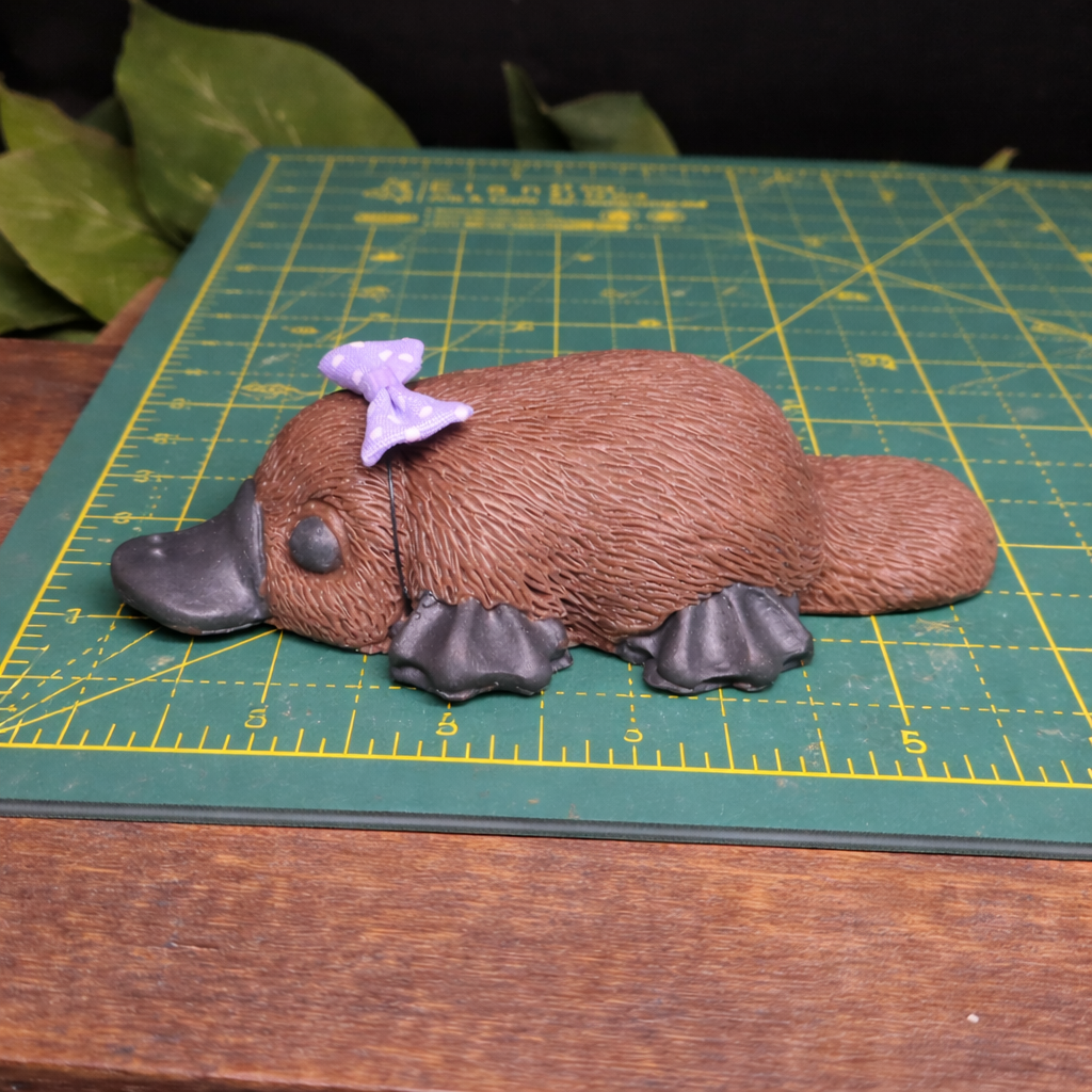 Handmade platypus goat milk soap with lavender bow shown on cutting mat for size reference, whimsical animal bath soap