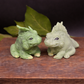 Two handmade iguana goat milk soaps in soft green tones posed facing each other on a neutral surface to show sculpted detail and scale.