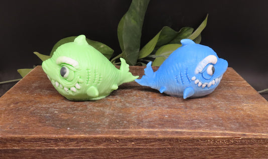 Two shark shaped soaps, one green and one blue, on a wooden surface with leaves in the background.